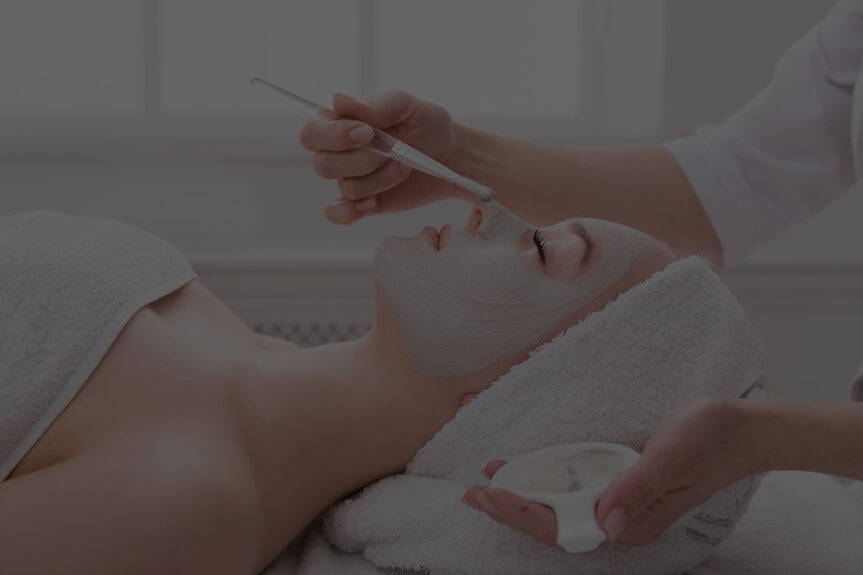 Person lying down with eyes closed, receiving a facial treatment with a brush applying cream.